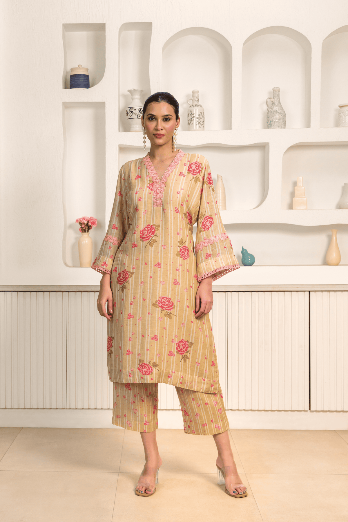 A model wearing a golden floral printed long tunic with matching cropped pants, paired with clear heels in a minimalist interior setting.