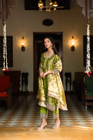 Side view of a woman in a traditional green anarkali outfit stands elegantly in a vintage-style room, surrounded by  decor and plush seating.