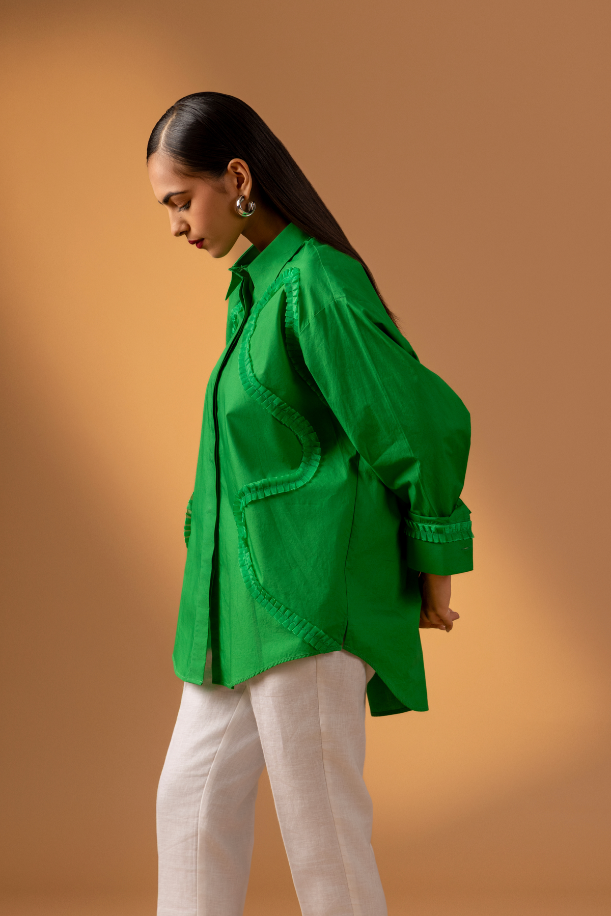 Side view of a model wears a vibrant green shirt with unique detailing, paired with white pants, against a soft brown background.