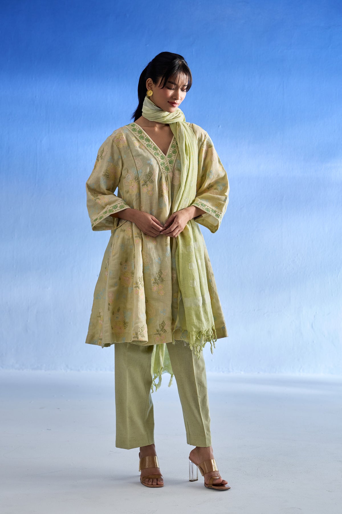 A woman modeling a pistachio green printed linen kurta set with matching trousers and a scarf against a blue gradient studio background.