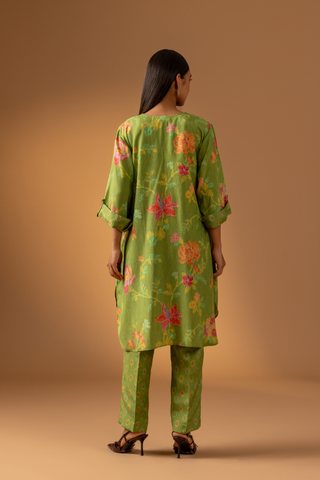 Back view of a model stands in a vibrant green co-ord set with floral patterns, paired with matching trousers, against a warm beige background.