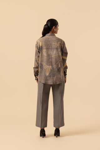 Back view of a model stands against a light beige background, wearing a patterned grey co-ord set which has button-up shirt and wide-legged beige pants, accessorized with heels.