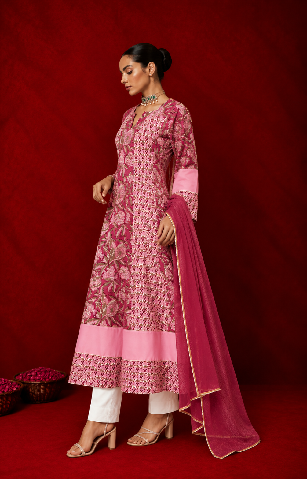 Left side of a woman wearing a pink floral printed anarkali kurta with white pants and a matching dupatta, styled with traditional jewelry, posing against a rich red backdrop.