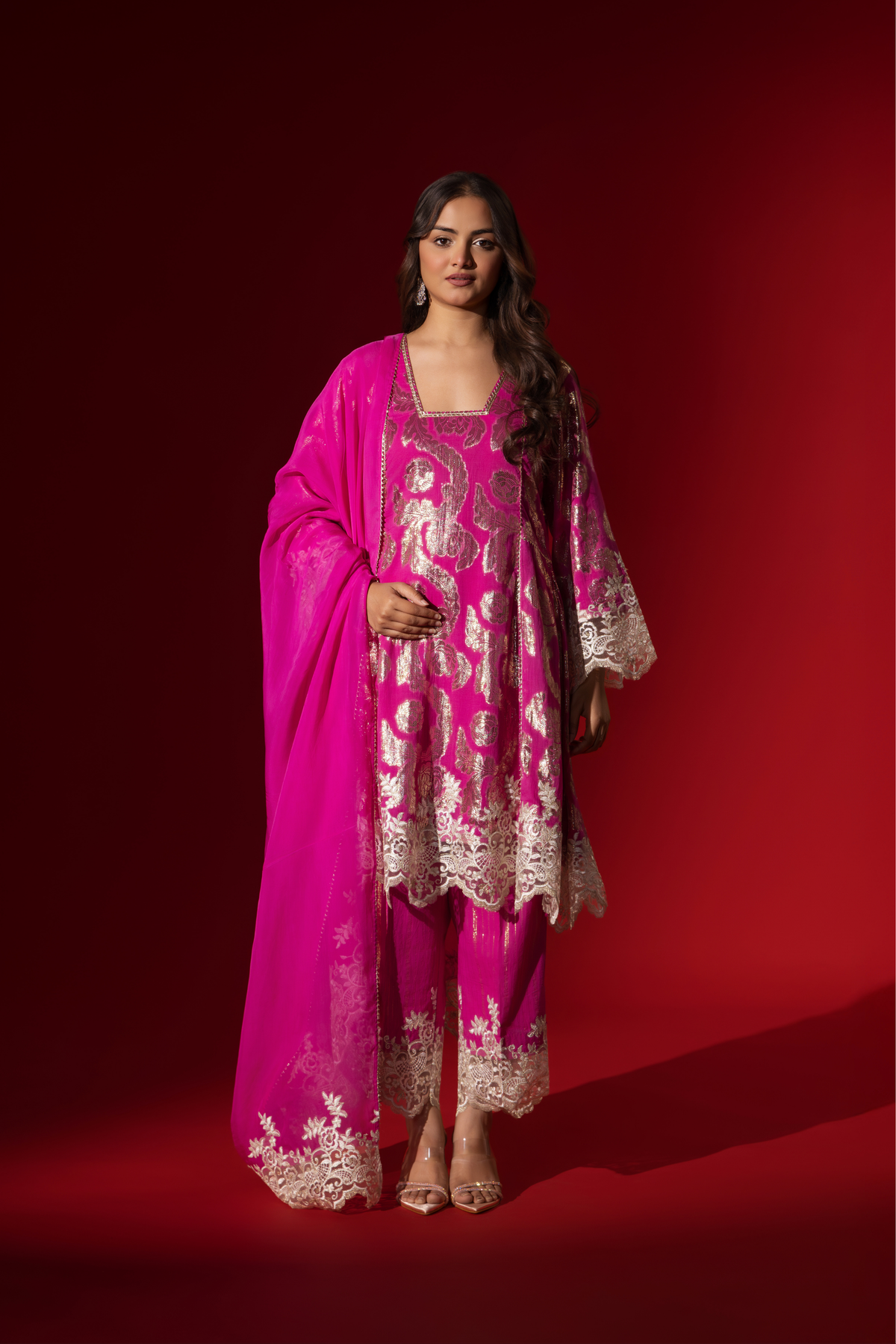 A woman in an elegant pink shimmer kurta set with intricate golden embroidery stands against a vibrant red background.