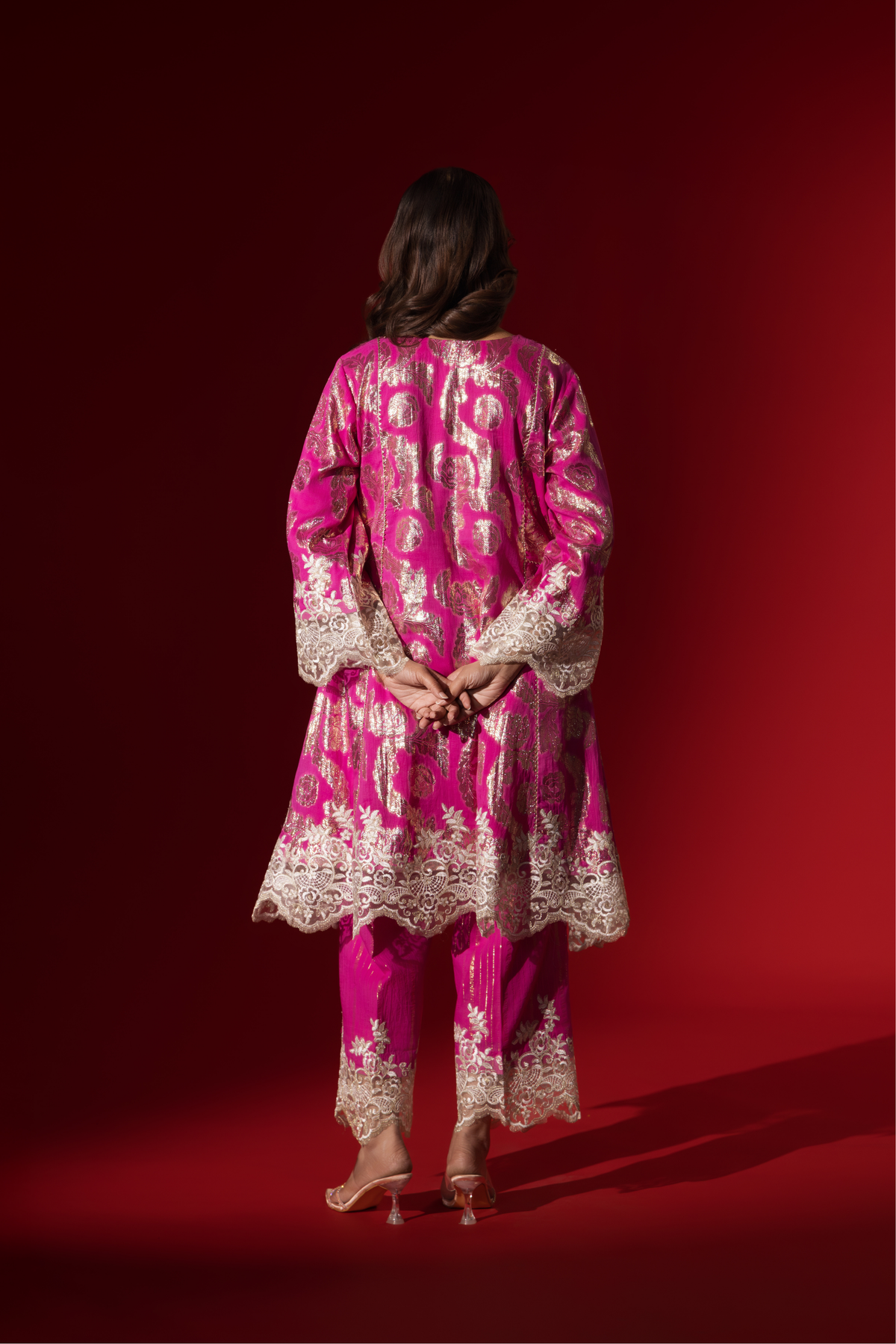 Back view of a woman in an elegant pink shimmer kurta set with intricate golden embroidery stands against a vibrant red background.