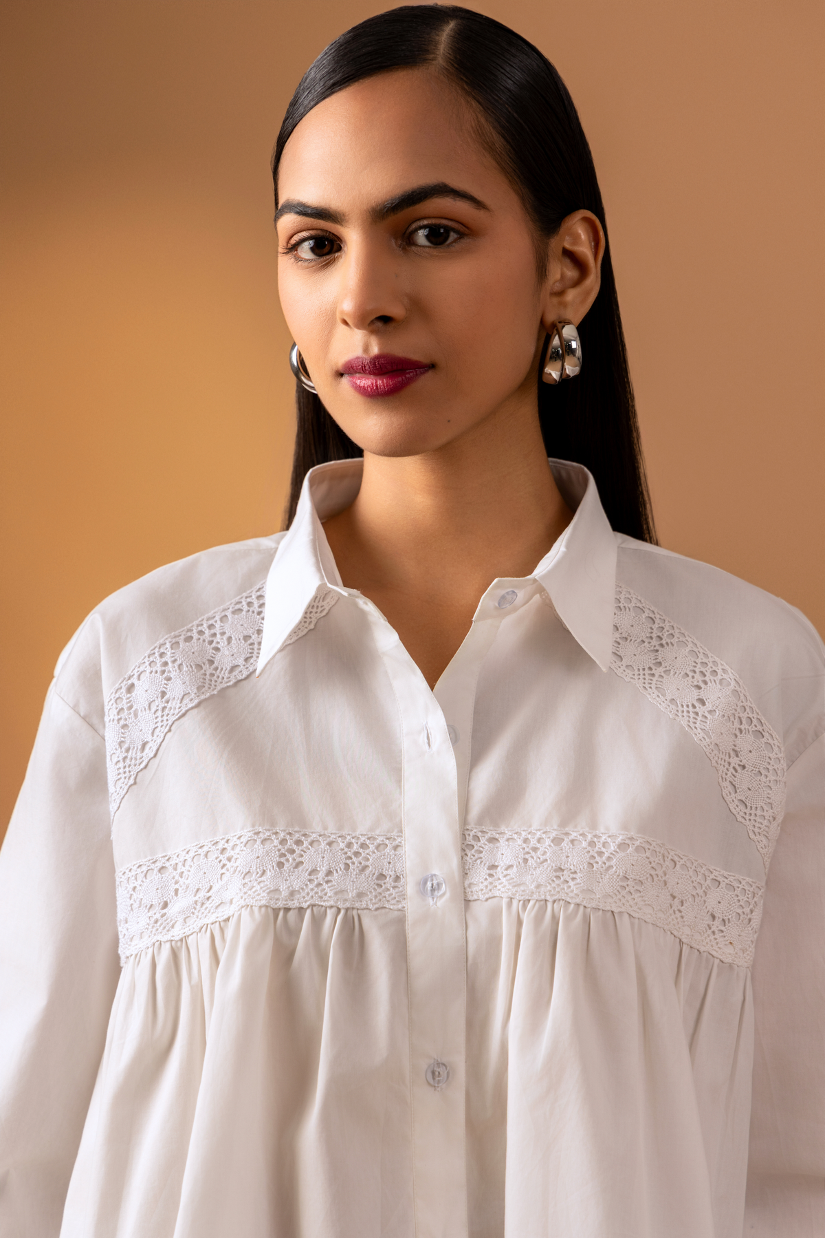 Closeup view of a model wearing a white button-down shirt with lace detailing, paired with blue jeans, set against a warm beige backdrop.