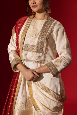A closup of a woman's hand showing traditional anarkali kurta set with gold embellishments and a red dupatta, posed against a rich red background.