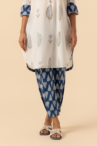 A person stands wearing a light-colored, floral-printed kurta paired with blue patterned pants and sandals against a neutral backdrop.