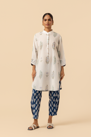 A woman stands wearing a light-colored, floral-printed kurta paired with blue patterned pants and sandals, against a beige background.