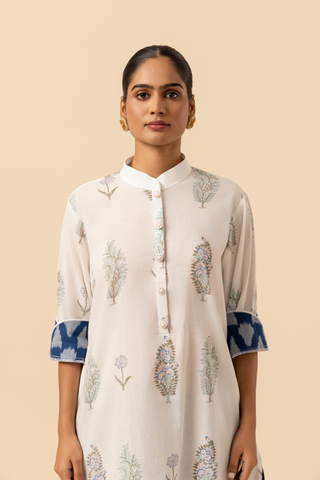 Closeup view of a woman stands wearing a light-colored, floral-printed kurta paired with blue patterned pants and sandals, against a beige background.