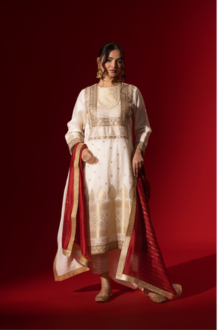 A woman in a white traditional outfit with gold embellishments and a red dupatta, posed against a rich red background.