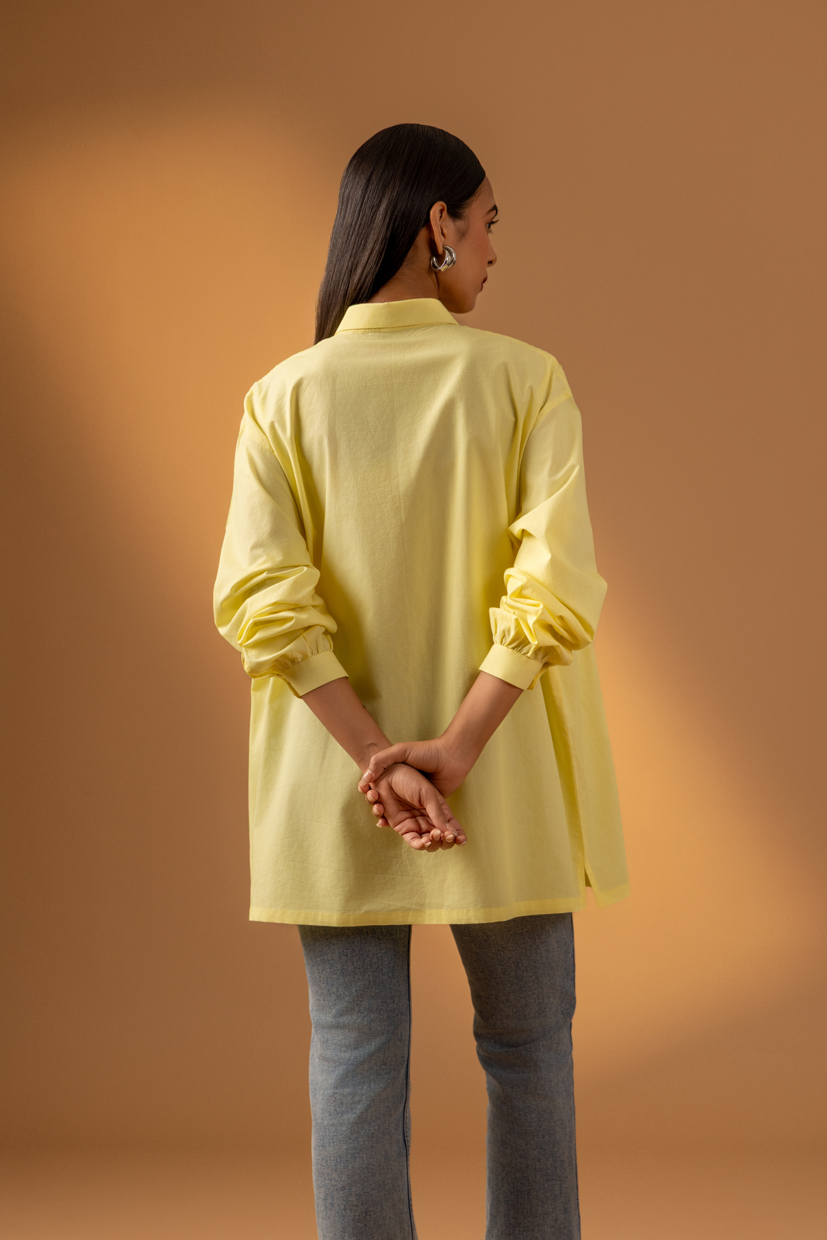 Side view of a woman wearing a stylish, oversized yellow shirt with delicate pleating, paired with light denim jeans, set against a warm backdrop.