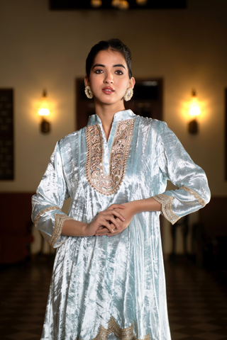 Closeup view of a woman wears a light blue ethnic velvet co-ord set outfit, standing gracefully in a spacious, elegantly decorated room with warm lighting and traditional furniture.