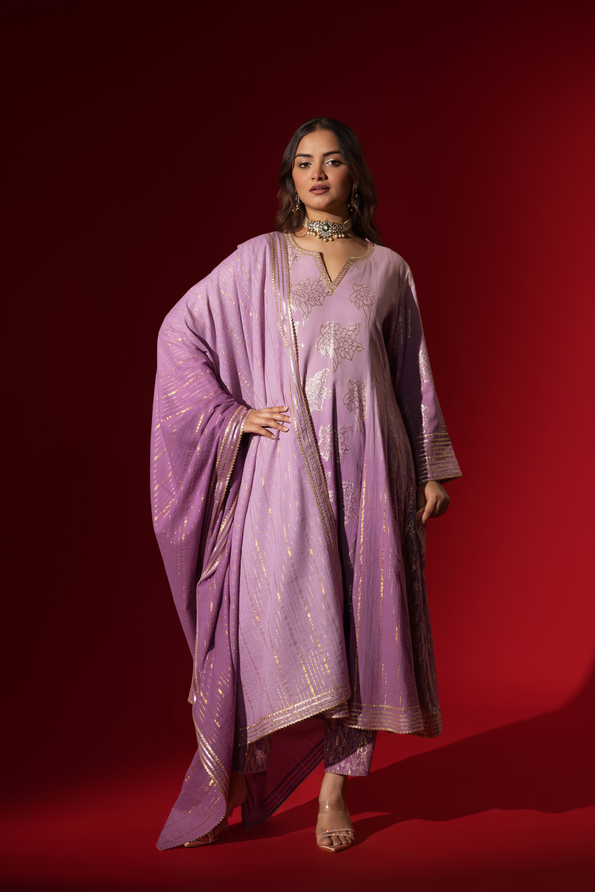 A woman showcases an elegant lilac anarkali kurta set with intricate designs, contrasting beautifully against a deep red background.