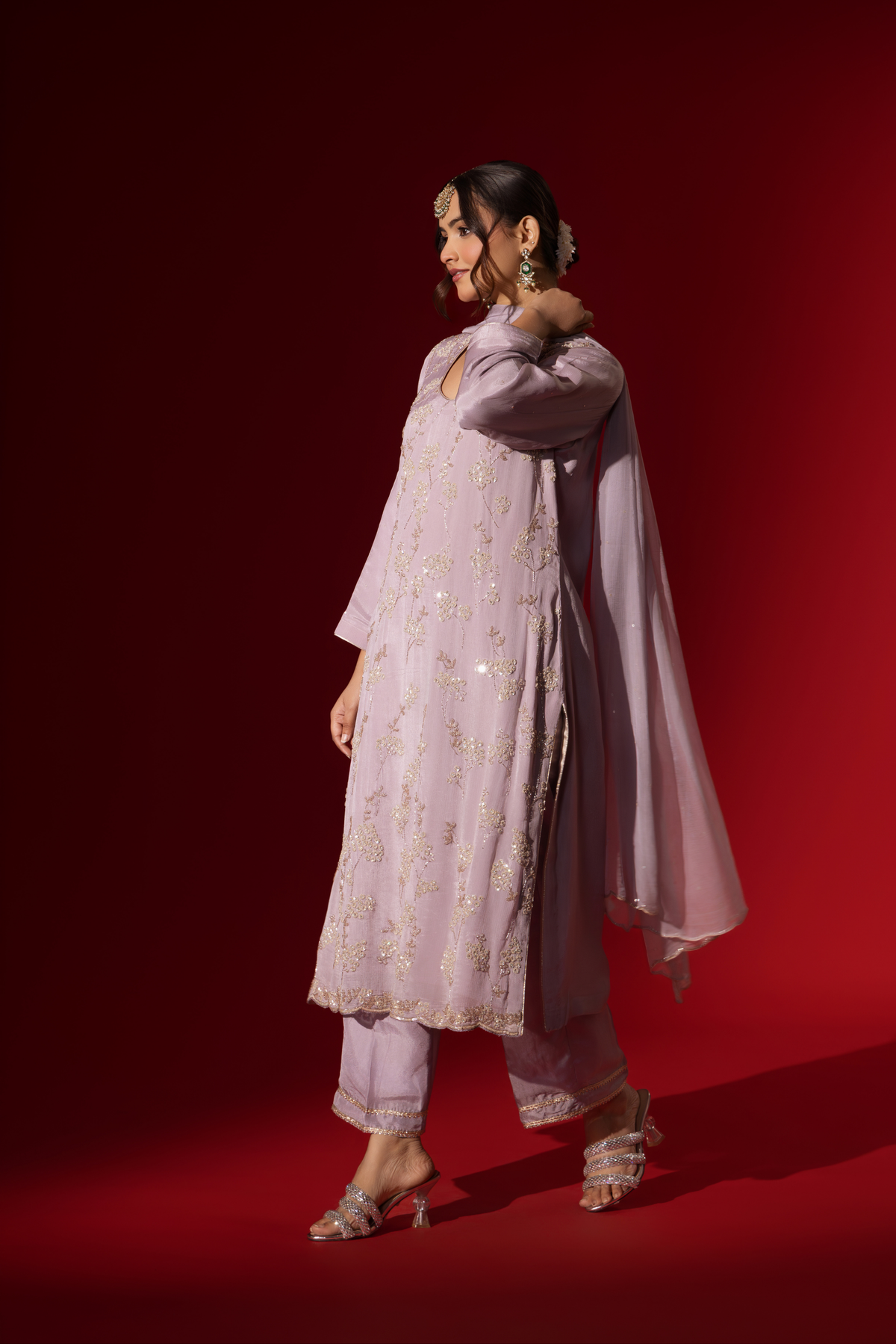 Side view of a woman dressed in a Lilac Radiance Embroidered Kurta Set stands confidently against a deep red background. Elegant traditional attire.