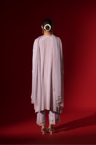 Back view of a woman dressed in a Lilac Radiance Embroidered Kurta Set stands confidently against a deep red background. Elegant traditional attire.