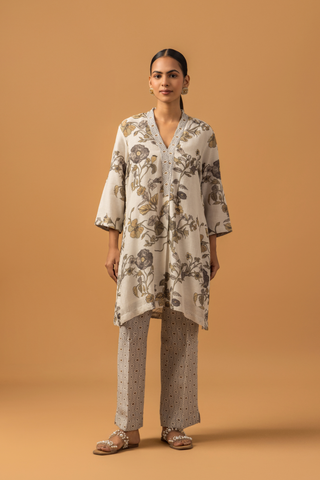 A woman stands against a warm tan background wearing a loose, floral-patterned co-ord set over patterned pants. The tone is calm and minimalist, with neutral colors 