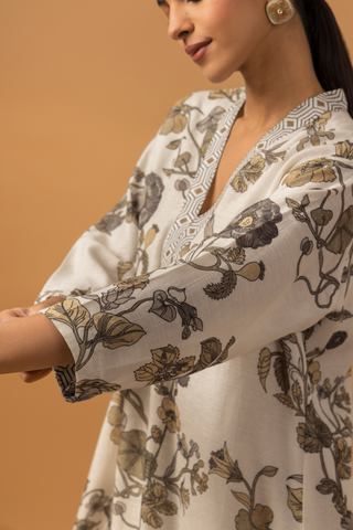 Closeup view of a woman stands against a warm tan background wearing a loose, floral-patterned co-ord set over patterned pants and showing her hand sleeves. 
