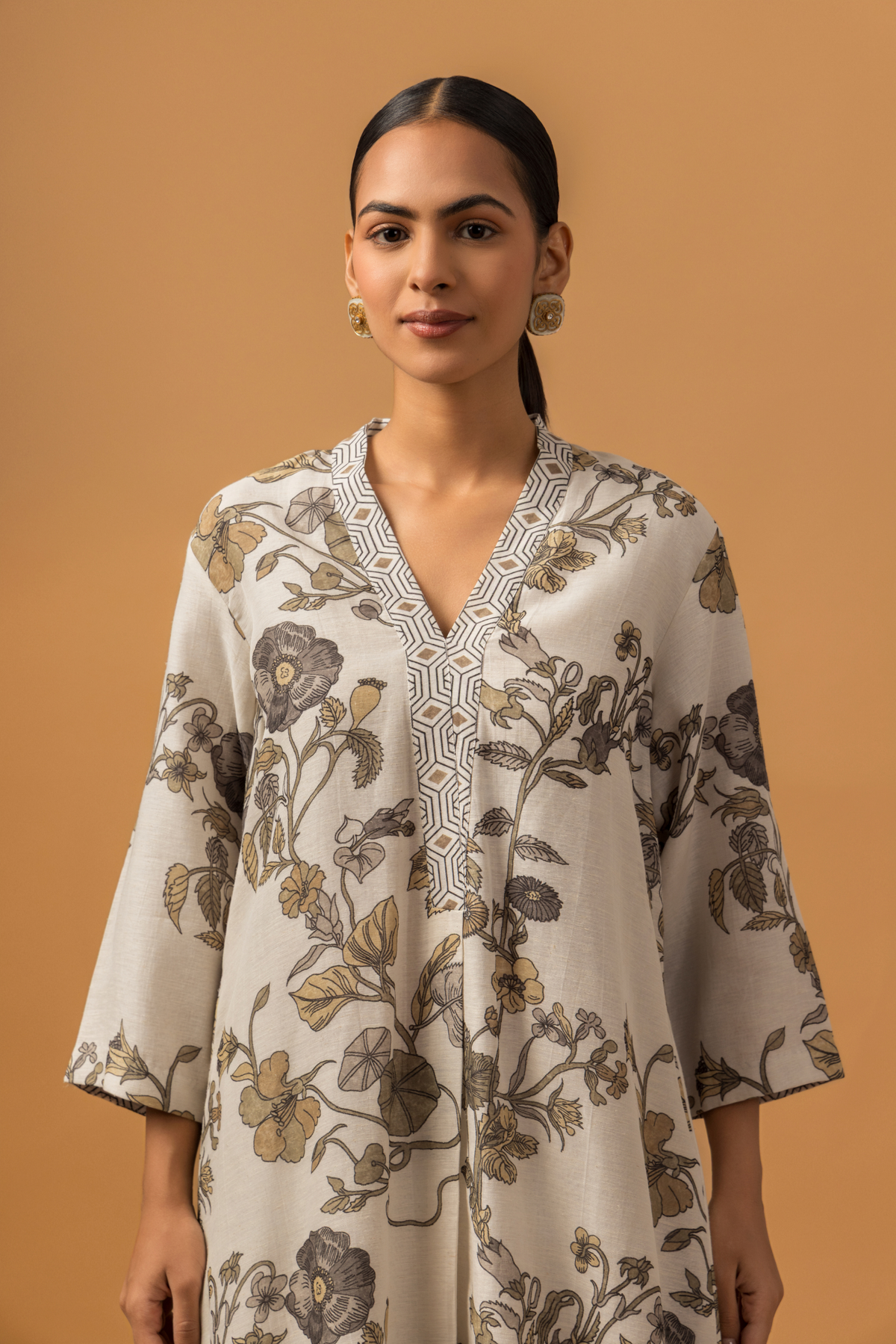 Closeup view of a woman stands against a warm tan background wearing a loose, floral-patterned co-ord set over patterned pants. The tone is calm and minimalist, with neutral colors 