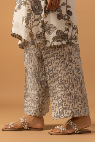 A person in patterned beige pants and a floral top stands on a brown background. They wear pearl-sandals, conveying a chic, casual style.