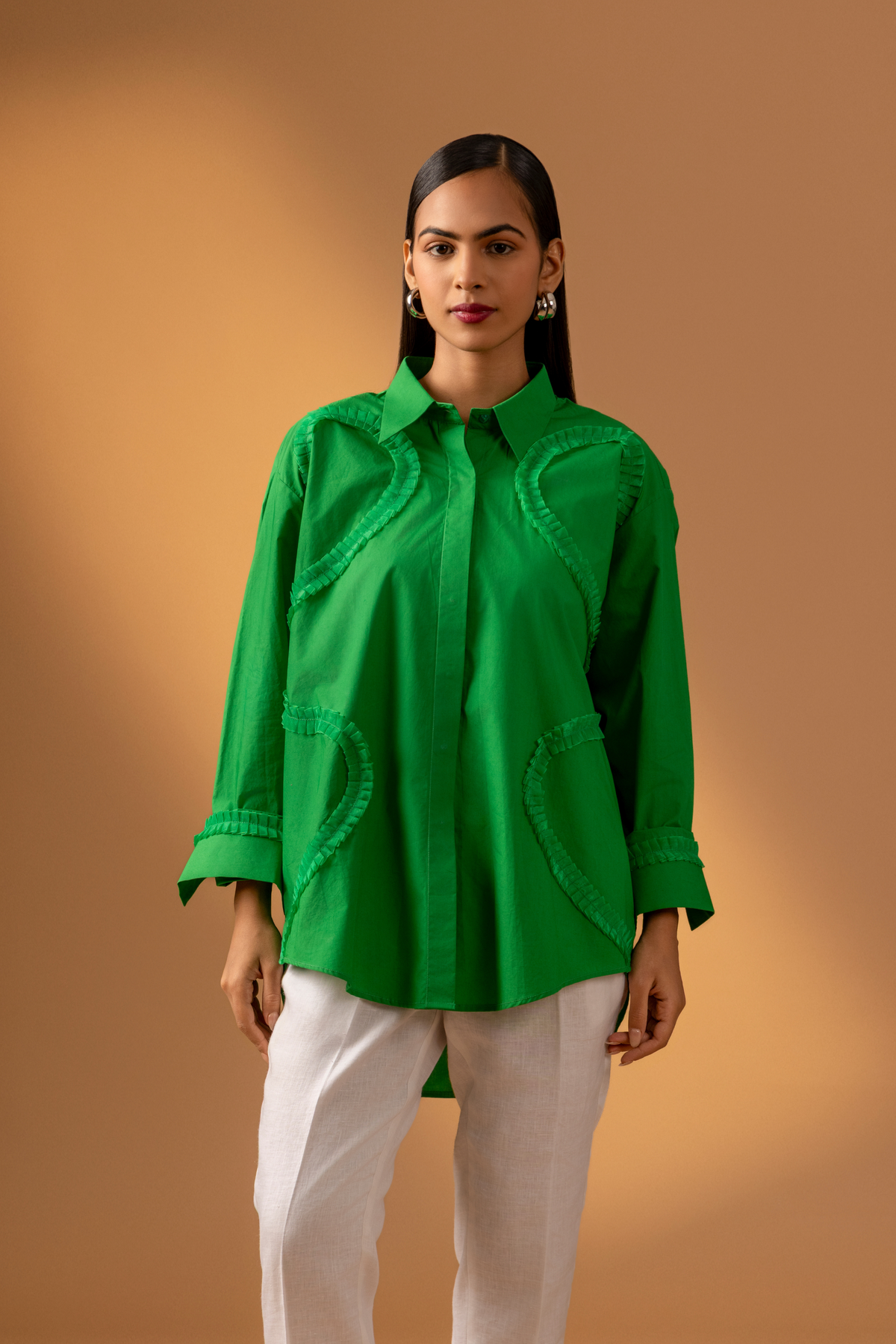 A model wears a vibrant green shirt with unique detailing, paired with white pants, against a soft brown background.
