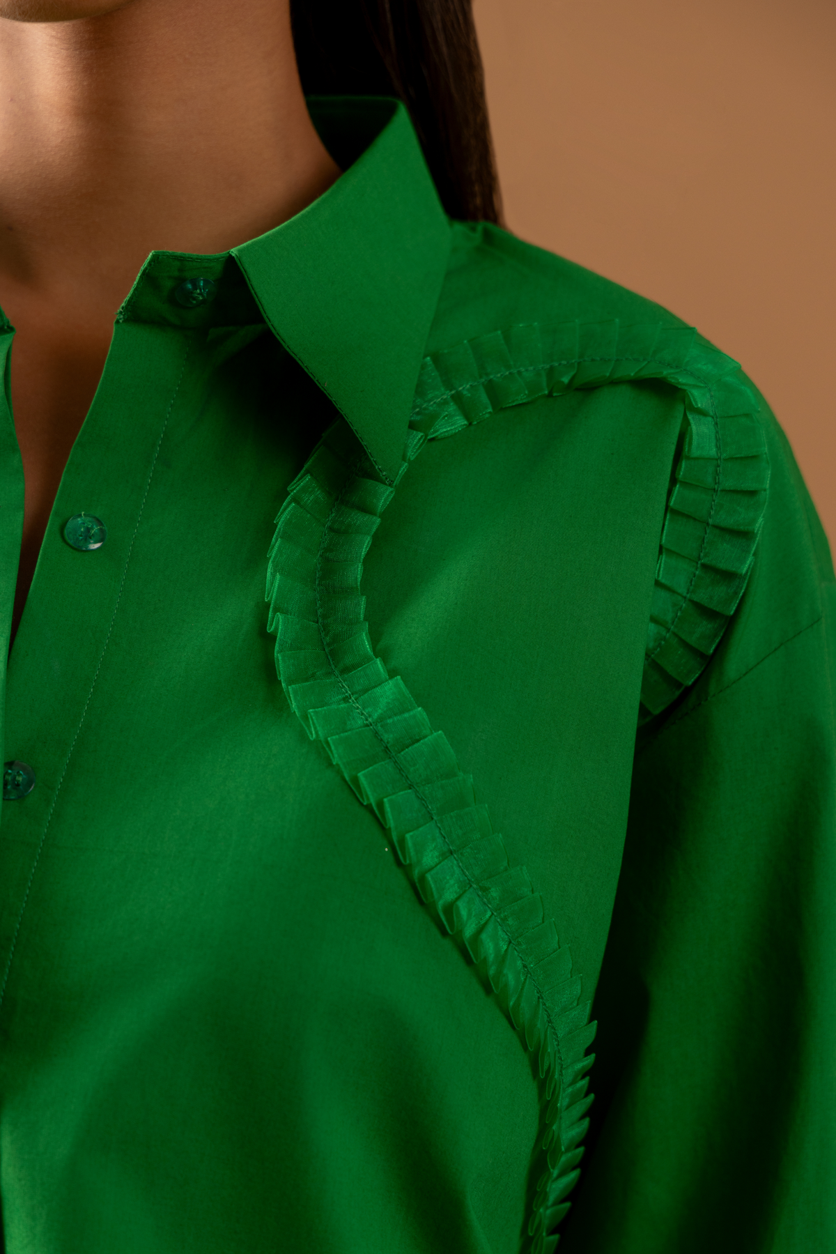 Closeup shirt view of a model wears a vibrant green shirt with unique detailing, paired with white pants, against a soft brown background.