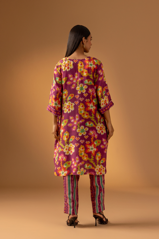 Back view of a model showcases a magenta floral-patterned co-ord set paired with colorful striped pants against a warm, neutral background.