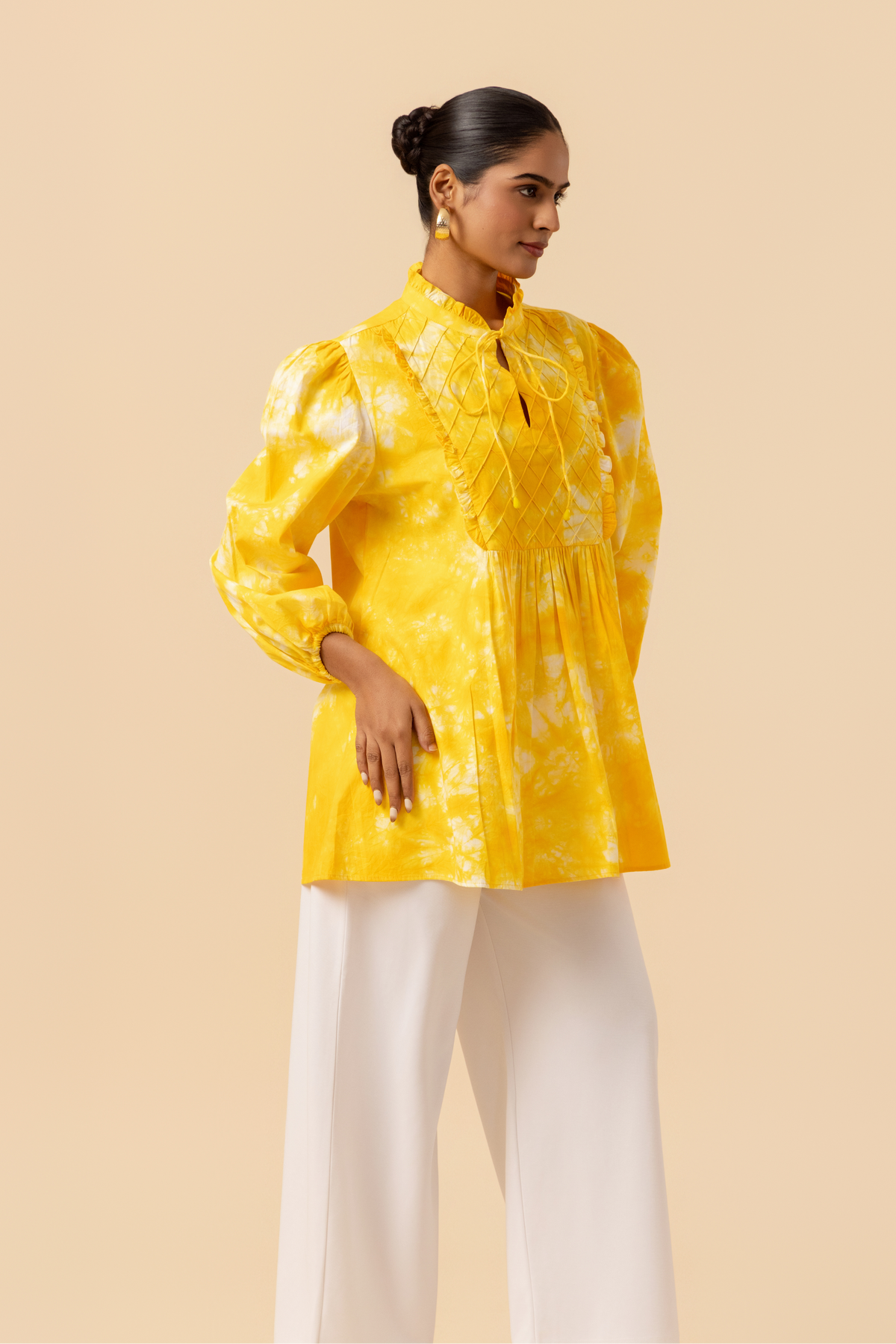 Side view of a woman wears a vibrant yellow tie-dye shirt dress with puffed sleeves, paired with white wide-leg trousers and stylish sandals.