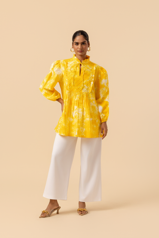 A woman wears a vibrant yellow tie-dye shirt dress with puffed sleeves, paired with white wide-leg trousers and stylish sandals.