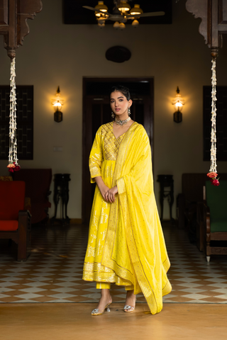 Side view of a  woman in a yellow anarkali kurta with intricate gold patterns with matching pants and dupattaa, standing in a beautifully decorated interior.