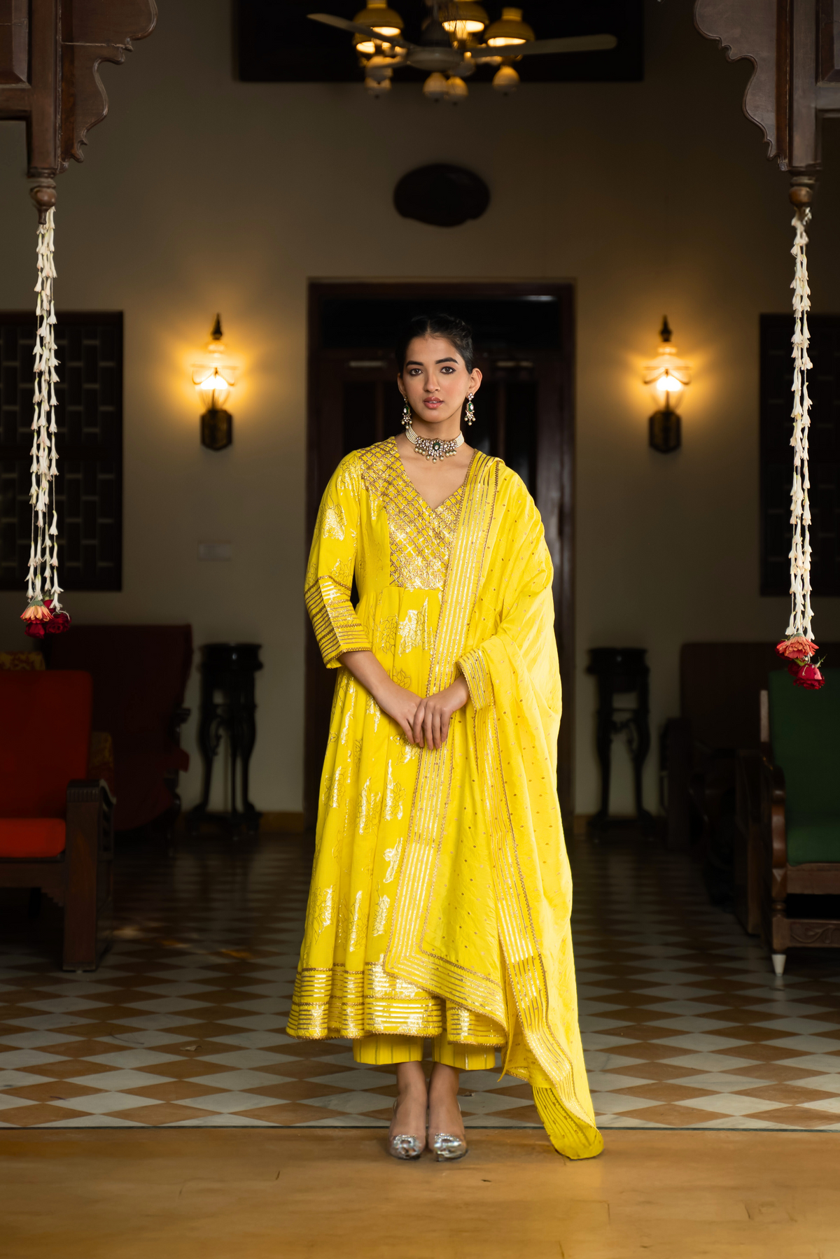 A woman in a yellow anarkali kurta with intricate gold patterns with matching pants and dupattaa, standing in a beautifully decorated interior.