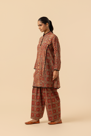 Side view of a model showcases a vibrant, patterned ethnic ajrakh printed farshi co-ord set featuring a long tunic and matching pants in rich red tones.