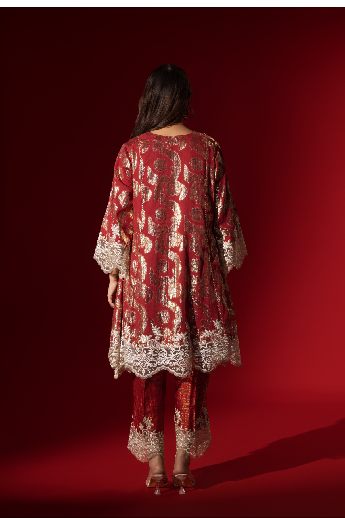 Back view of a woman poses in a maroon ethnic  shimmer kurta set outfit with intricate white embroidery against a vibrant red backdrop.