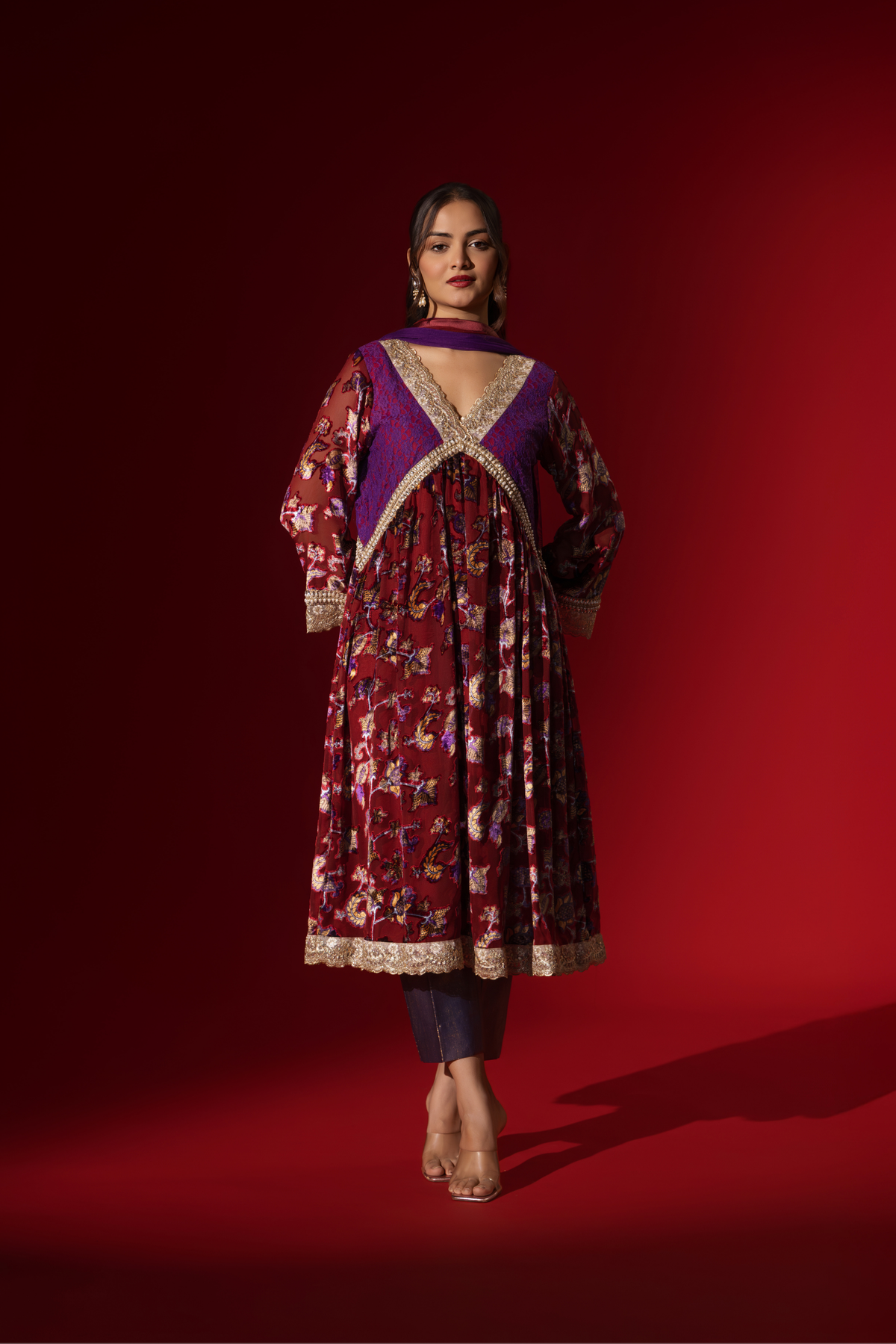 A model poses in a vibrant maroon floral velvet anarkali kurta set with purple accents and a golden trim, set against a rich red background.