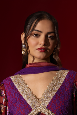 Closeup view of a model poses in a vibrant maroon floral velvet anarkali kurta set with purple accents and a golden trim, set against a rich red background.