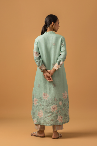 Back view of a woman in a pastel green, floral co-ord set with delicate embroidery stands against a beige background. She is wearing beige pants, sandals, and hoop earrings, conveying a serene and elegant tone.