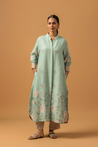 A woman in a pastel green, floral co-ord set with delicate embroidery stands against a beige background. She is wearing beige pants, sandals, and hoop earrings, conveying a serene and elegant tone.