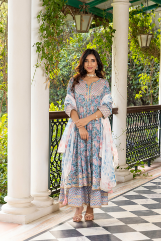 A woman stands elegantly in a beautiful, patterned block printed ethnic kurta set with intricate details, surrounded by lush greenery and architectural columns.