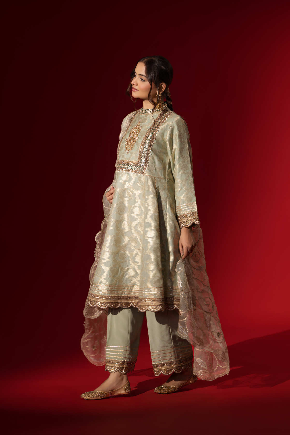 Side view of a woman wearing an elegant mint green embroidered sequin brocade kurta set with gold accents and a sheer dupatta, set against a rich red background
