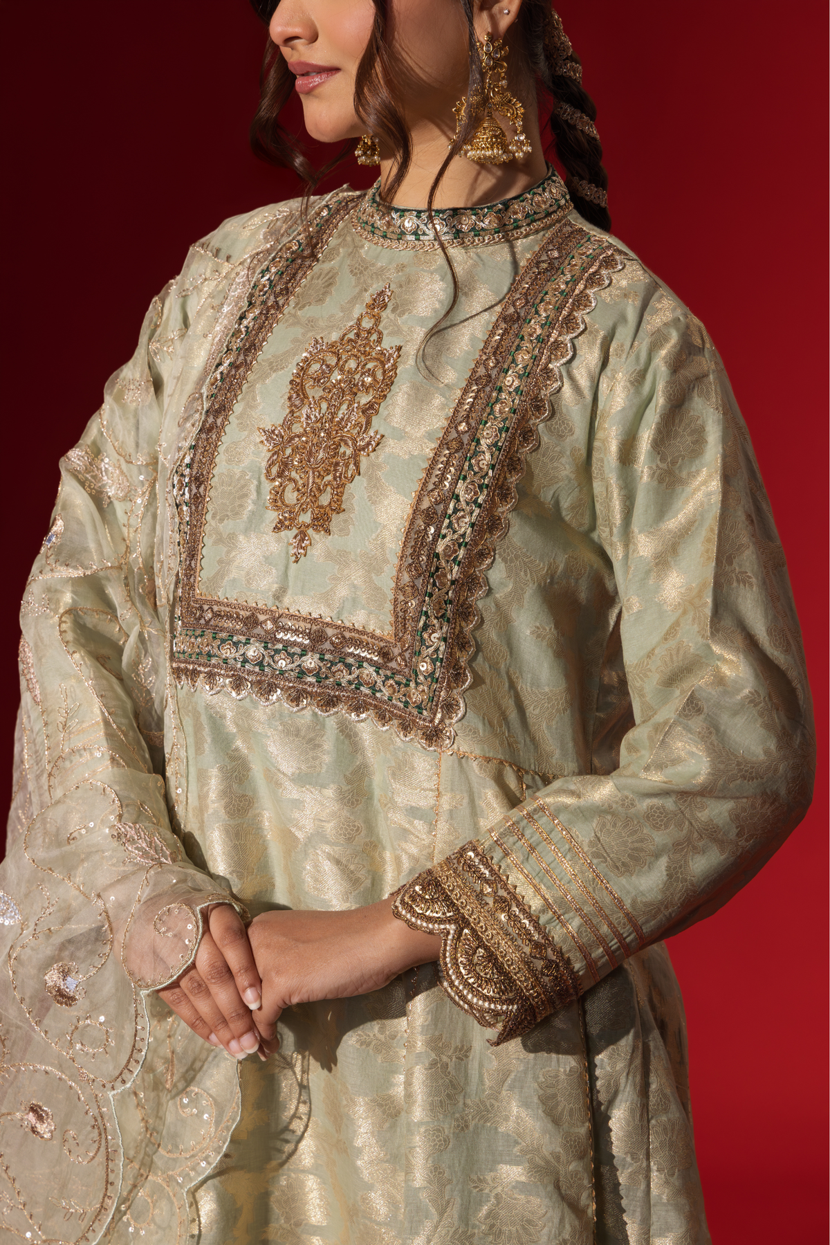 Closeup view of a woman's hand wearing an elegant mint green embroidered sequin brocade kurta set with gold accents and a sheer dupatta, set against a rich red background