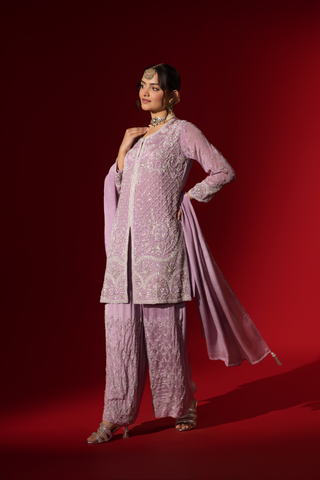 Side view of a woman stands confidently in a beautifully lavender embroidered kurta set, against a rich red background.