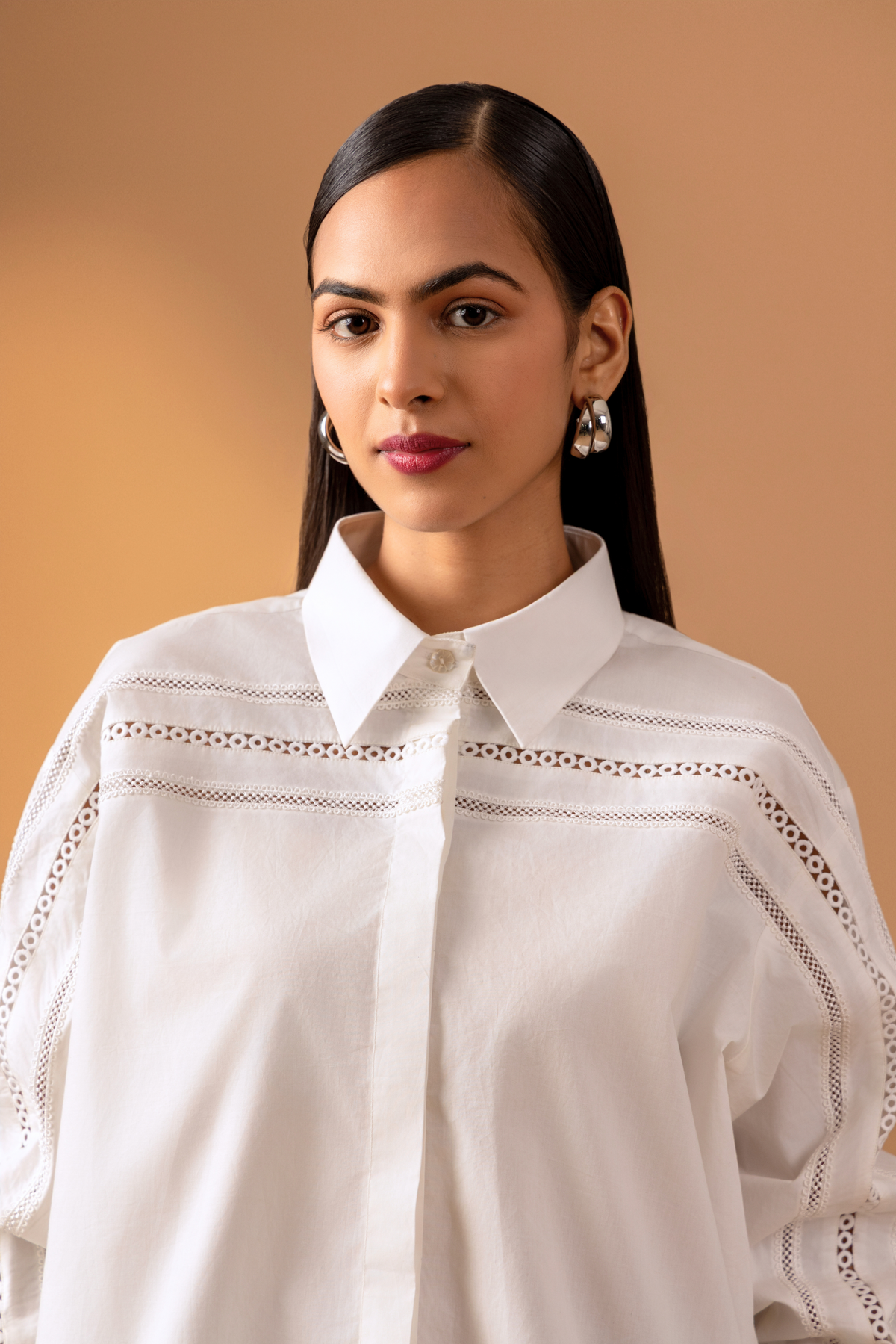 Close-up view of a women stands confidently in a stylish white button-up shirt with intricate details and puffed sleeves, set against a warm beige background.