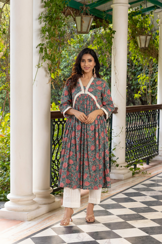 A woman stands elegantly on a black-and-white checkered floor, wearing a floral handblock printed cotton kurta set and white pants, surrounded by greenery.