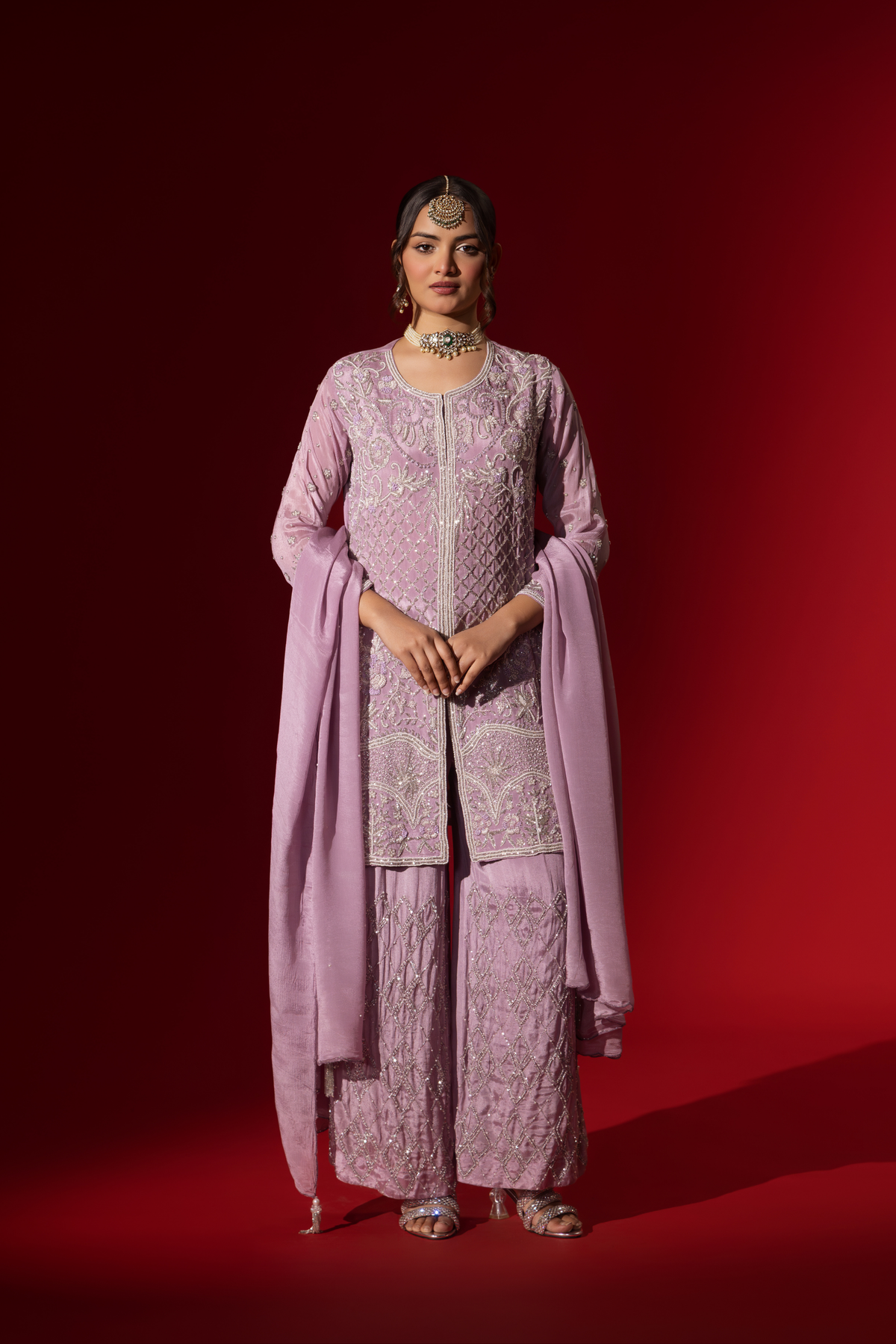 A person stands confidently in a beautifully lavender embroidered kurta set, against a rich red background.