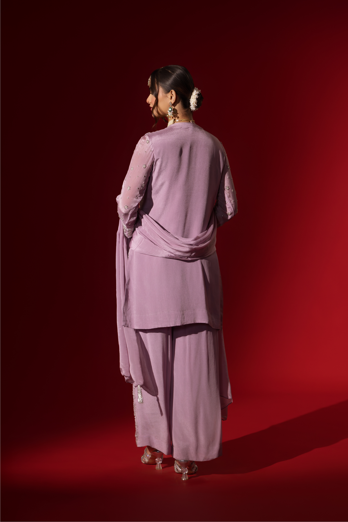 Back view of a woman stands confidently in a beautifully lavender embroidered kurta set, against a rich red background.