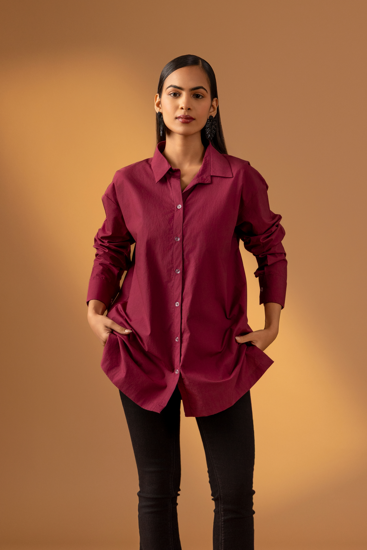 A woman wearing a maroon shirt with long sleeves paired with black pants against a warm, muted background, showcasing a stylish, casual look. 