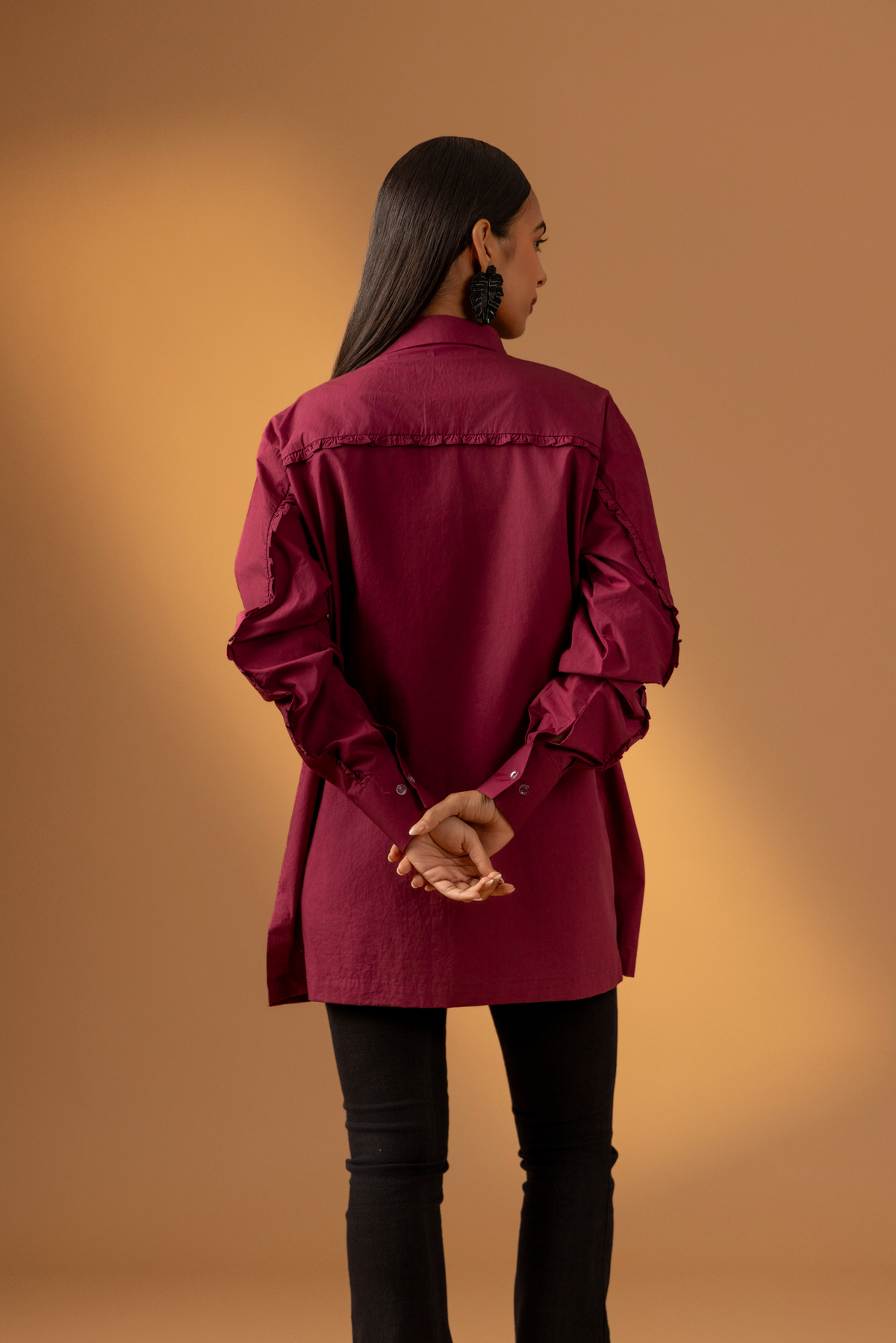 Back view of a woman wearing a maroon shirt with long sleeves paired with black pants against a warm, muted background, showcasing a stylish, casual look. 
