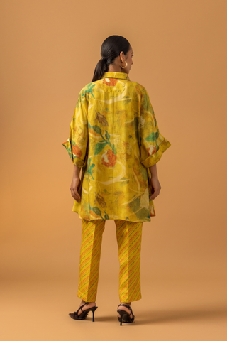 Back view of a model stands in a bright yellow co-ord set featuring a patterned oversized shirt and matching striped pants against a warm backdrop.