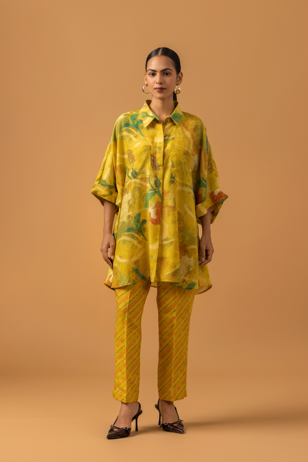 A model stands in a bright yellow co-ord set featuring a patterned oversized shirt and matching striped pants against a warm backdrop.