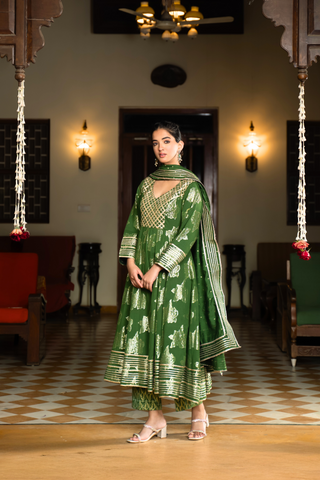 Side view of a woman in a vibrant green anarkali kurta set with matching pants stands gracefully in a well-decorated interior.